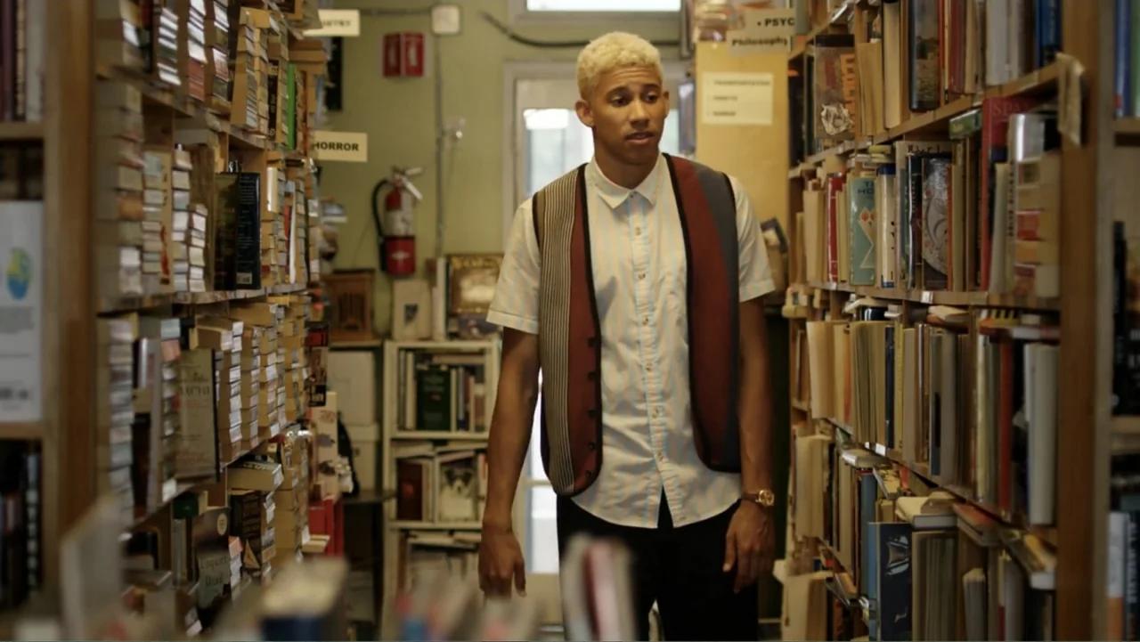 Like.Share.Follow. (2017) movie still: medium — A narrow aisle filled with wooden shelves packed tightly with various books. A young man stands…