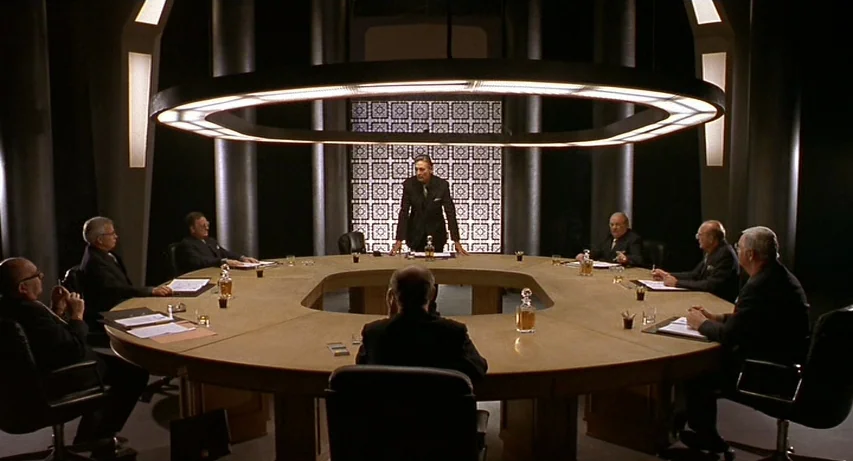 CQ (2001) movie still: wide — A dark, circular room contains a large wooden table under a ring-shaped light. Nine men in business suits occupy…