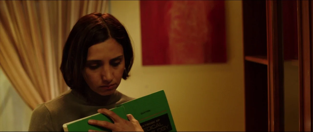 Under The Shadow (2016) movie still: medium — A dimly lit room with yellow walls and a large red rectangular art piece. A young woman holds a green…