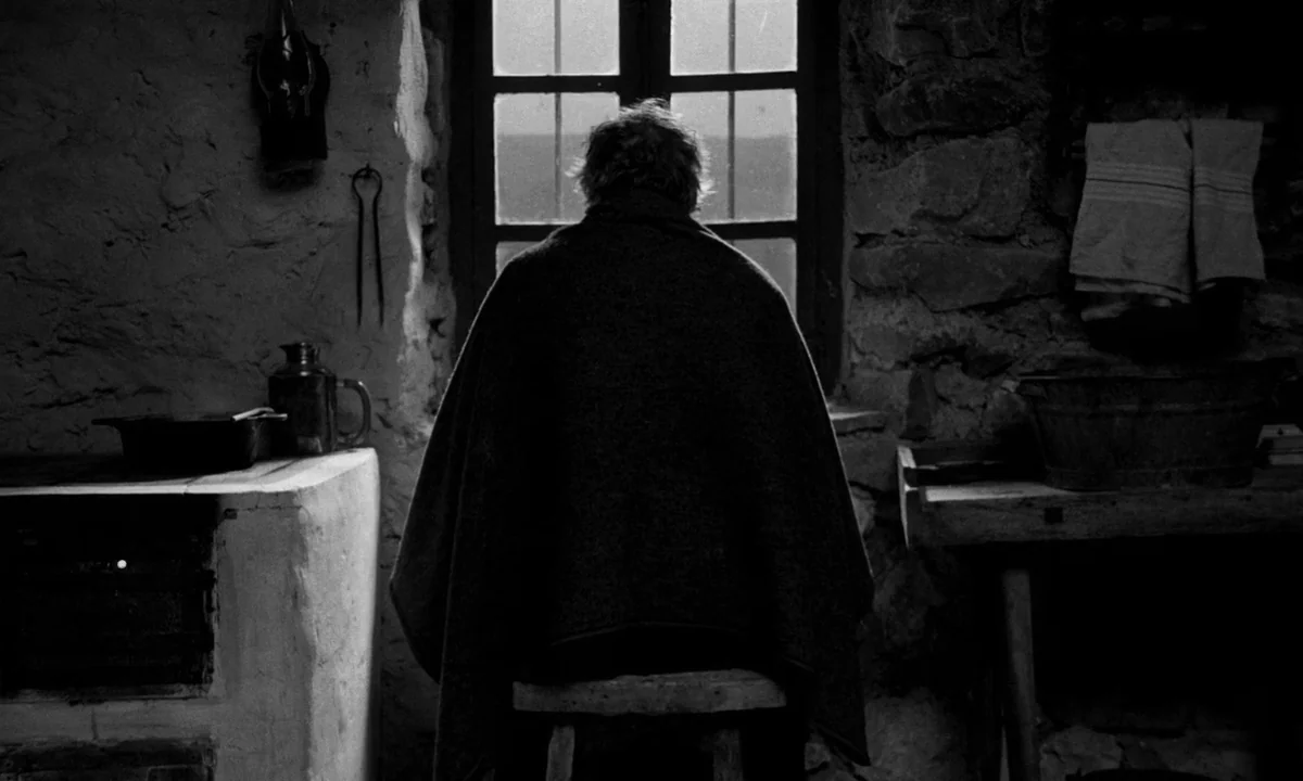 The Turin Horse (2011) movie still: medium — A rustic interior with thick stone walls and a small window in the center. An elderly person wrapped in…