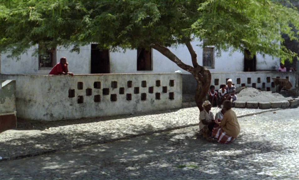 Casa De Lava (1994) movie still: wide — A stone building with a decorative block wall stands under a large leafy tree. People rest on the ground in…