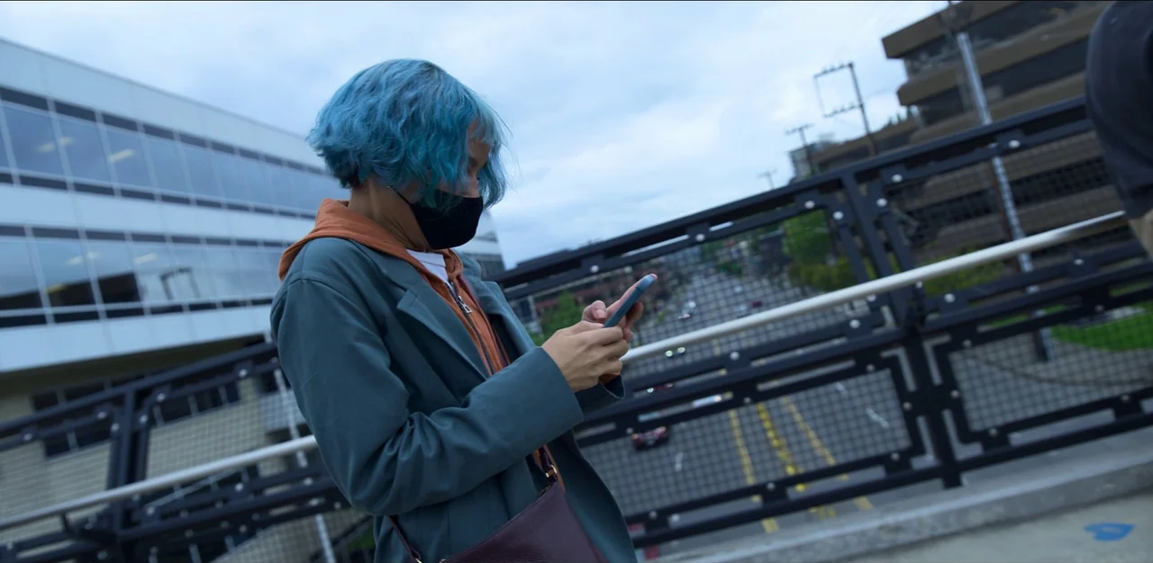 Kimi (2022) movie still: medium — A person with blue hair stands on a modern pedestrian bridge over a highway. The subject looks down at a…