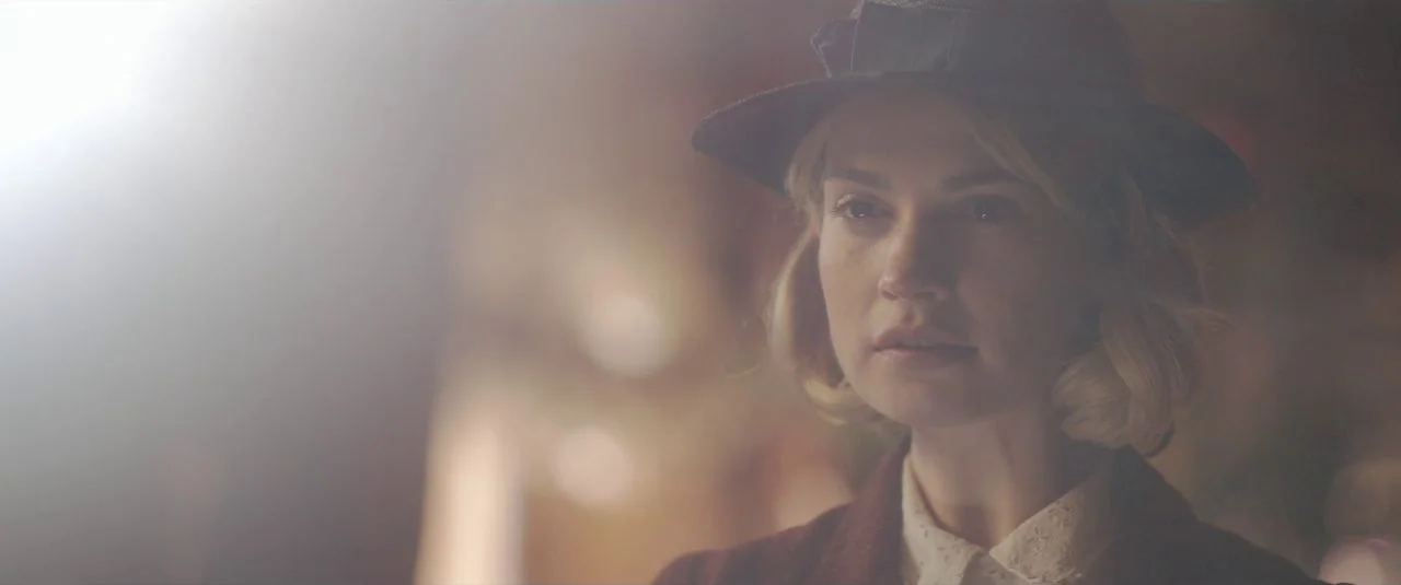 Rebecca movie still: closeup — A woman stands in a dimly lit, blurred indoor environment with a warm light source. She is wearing a…
