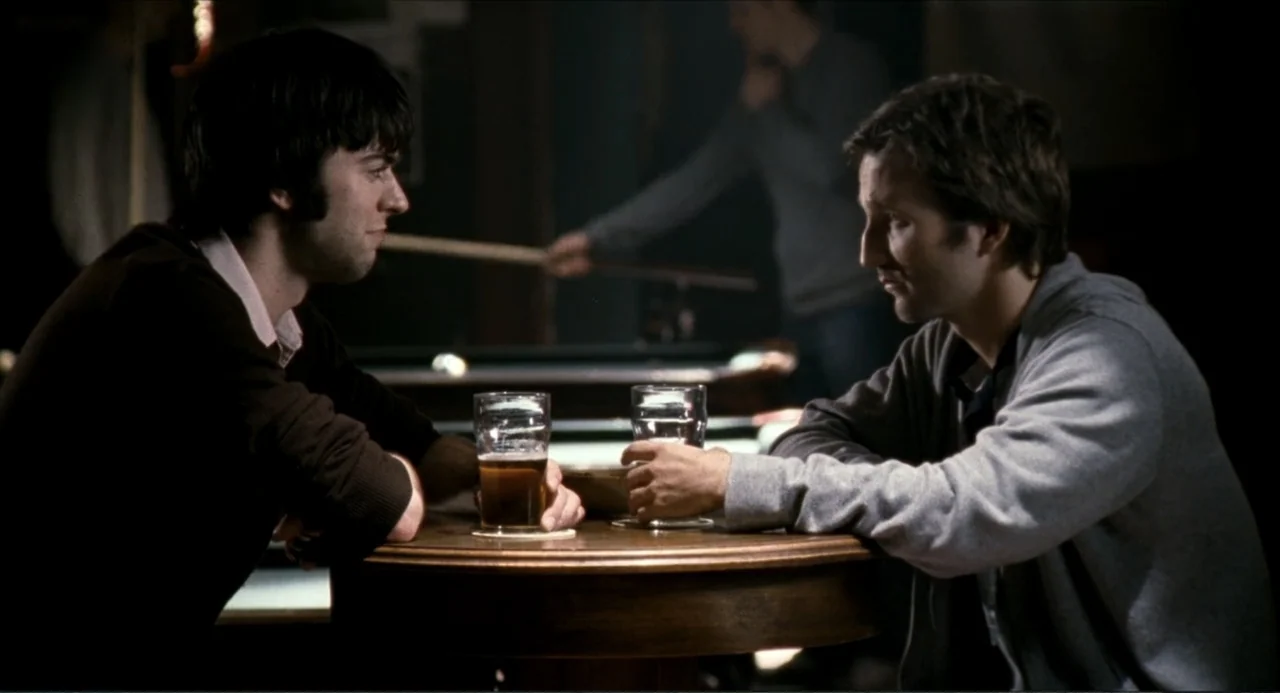 Colin Quinn: Red State, Blue State (2019) movie still: medium — A dark, dimly lit pub features a wooden circular table in the foreground. Two men…