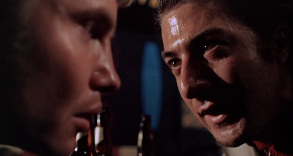 Midnight Cowboy (1969) movie still: closeup — Two men stand close together in a dimly lit bar interior. The men maintain eye contact as they speak…