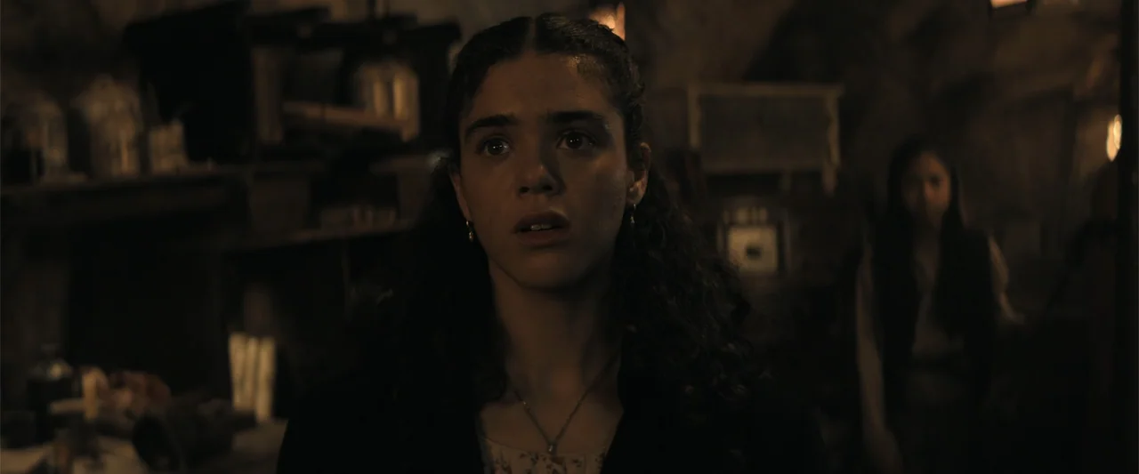 The Bluff (2026) movie still: medium — A dimly lit, cluttered workspace filled with shadows and indistinct objects in the background. A young woman…