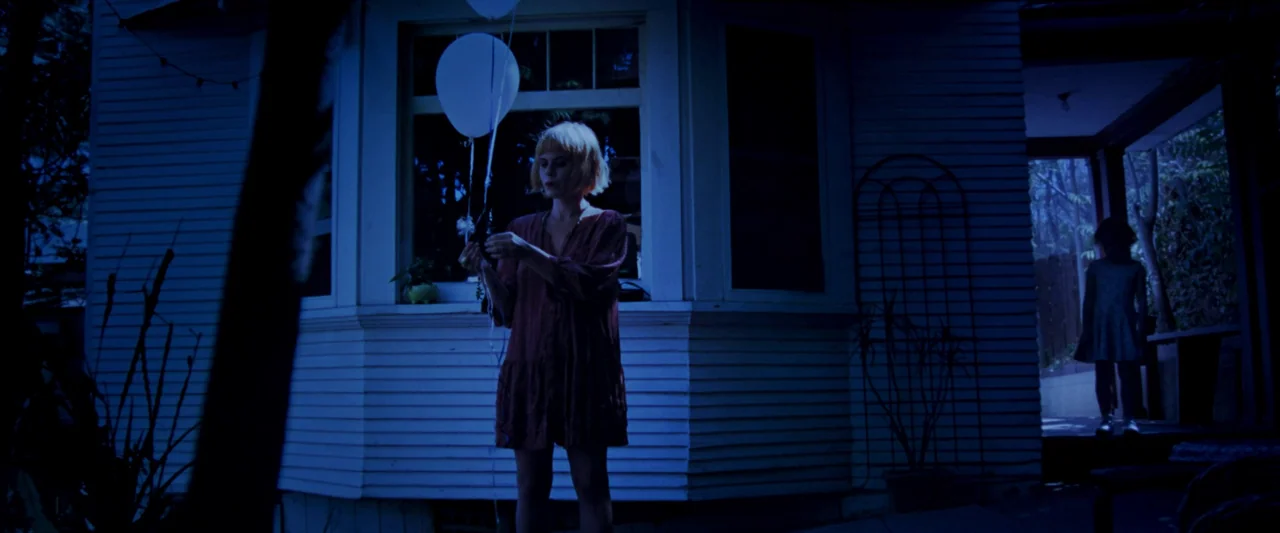 Moon Garden (2022) movie still: fullbody — A dark house exterior at night with horizontal white siding and a large window. A woman stands in the…