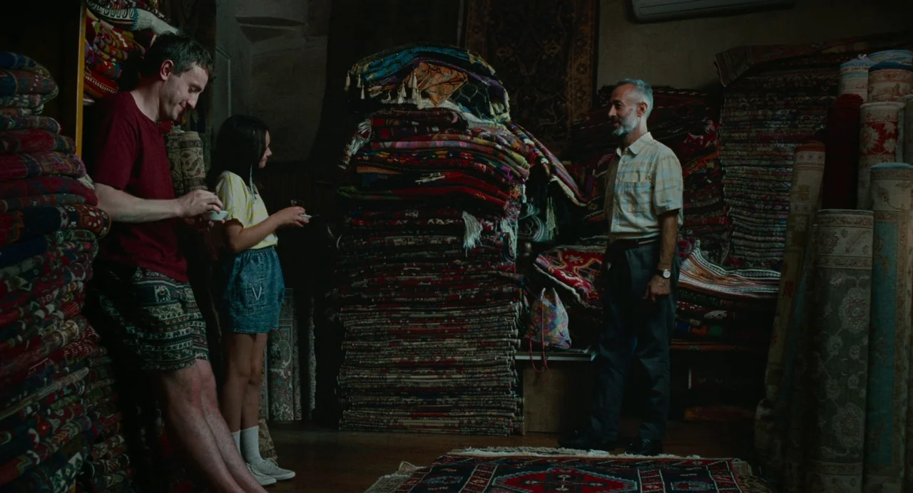 Aftersun (2022) movie still: wide — A dimly lit shop interior is filled with towering, dense stacks of intricate hand-woven rugs. A man and a girl…