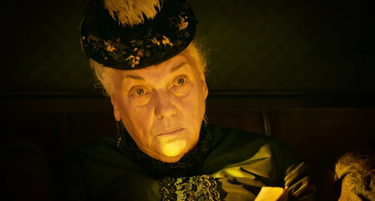 The Ballad of Buster Scruggs (2018) movie still: closeup — An elderly woman sits in a dimly lit room with ornate Victorian wall textures. She reads…