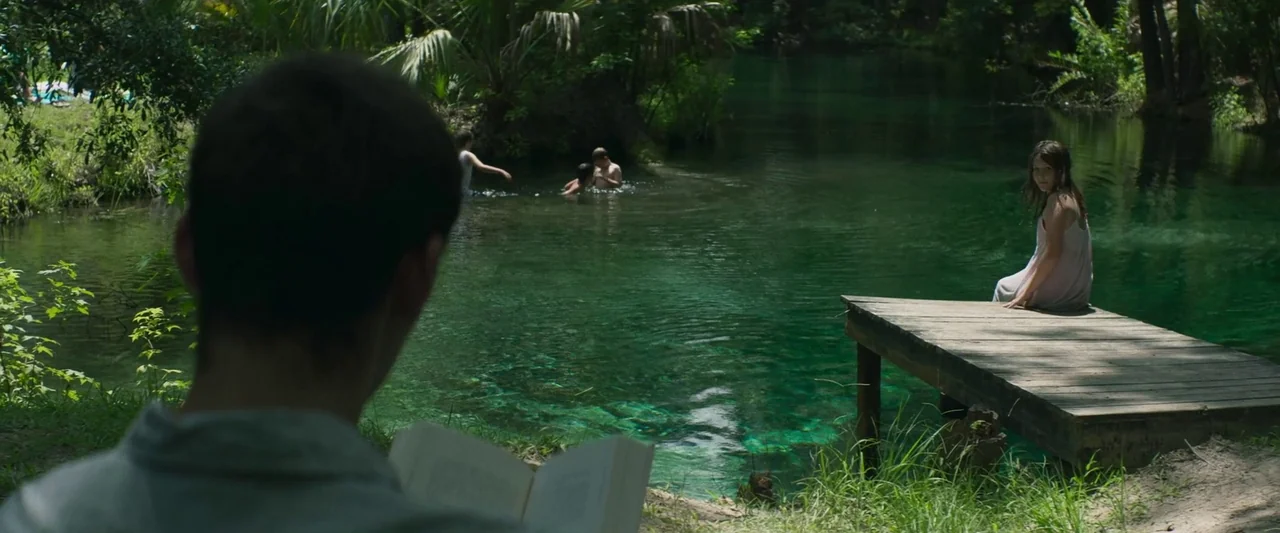 The Mad Hatter (2021) movie still: medium — A secluded natural swimming hole is surrounded by lush green vegetation and palm trees. A young woman…