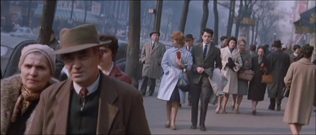Une Femme Est Une Femme (A Woman Is a Woman) (1961) movie still: medium — A crowded city sidewalk lined with mature trees features pedestrians…