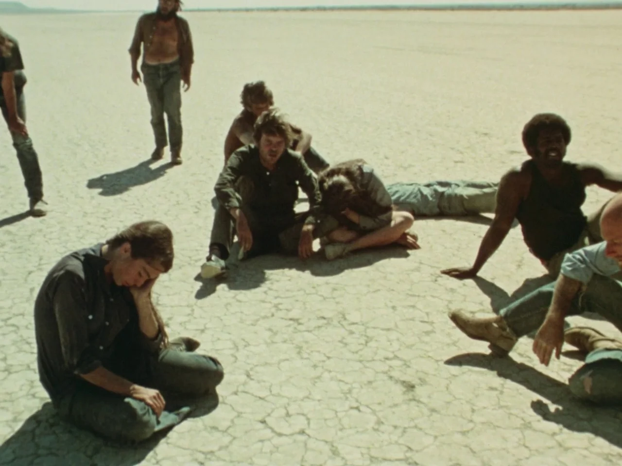 Punishment Park (1971) movie still: wide — A group of people sits scattered across a vast, cracked, dry desert landscape. The individuals rest in…