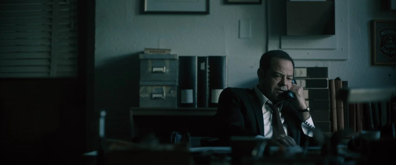 Boston Strangler (2023) movie still: medium — A dimly lit room contains stacked cardboard boxes and black binders on a desk. A man in a suit sits…