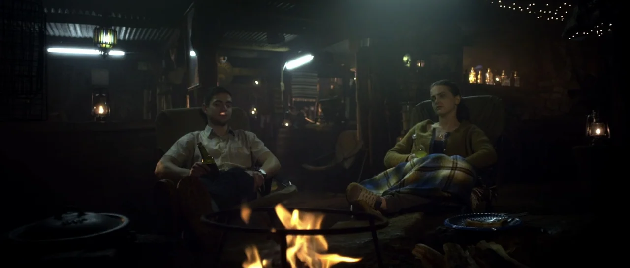 The Lullaby (2019) movie still: medium — A dimly lit cabin interior features a central fire pit with two people seated nearby. A man and a woman sit…