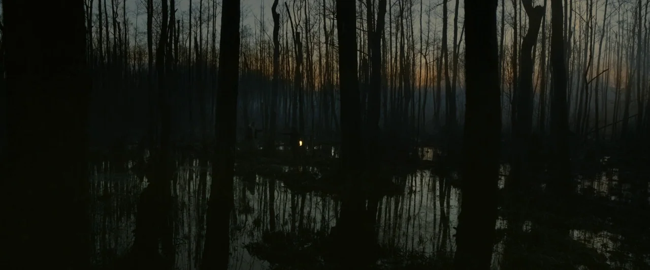 Vesper (2022) movie still: wide — A dense swamp filled with bare trees stands under a dim twilight sky. Dark water covers the ground reflecting the…