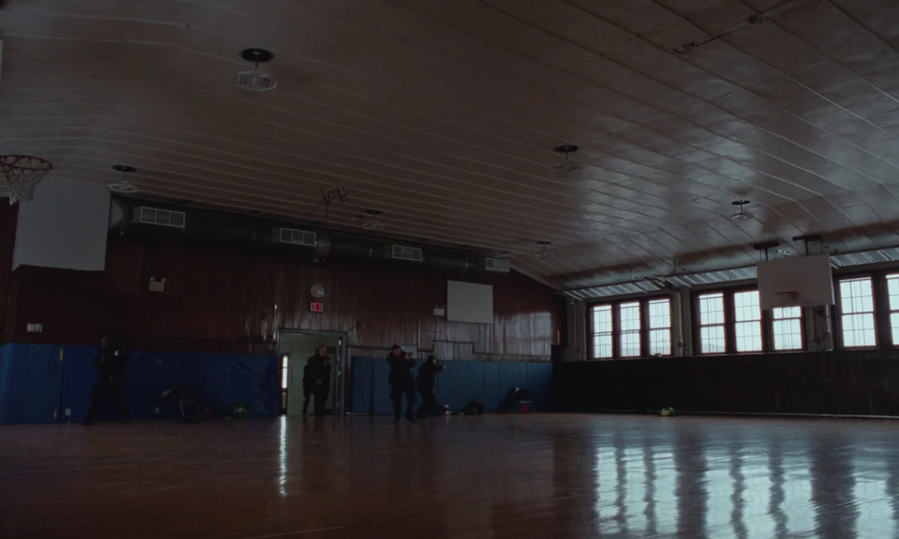 Vox Lux (2018) movie still: wide — A dark, expansive indoor gymnasium features polished wooden floors and wood-paneled walls. Several tactical…