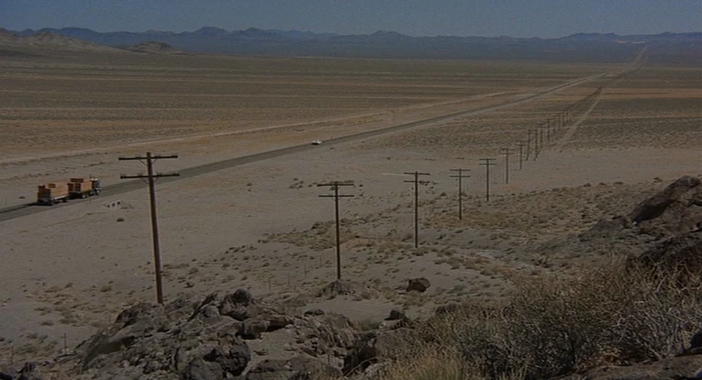 Vanishing Point (1971) movie still: establishing — A vast, arid desert expanse features a single highway cutting through the barren landscape. A…