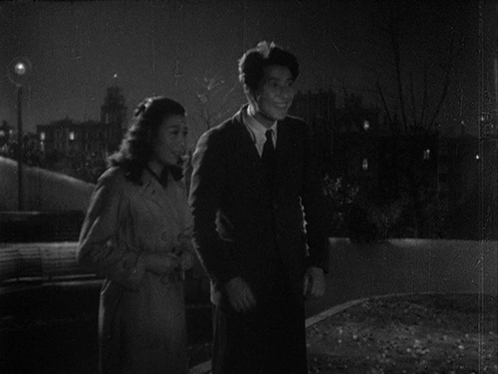 One Wonderful Sunday (1947) movie still: medium — A dark city park at night with blurred urban structures in the background. A man in a dark suit…