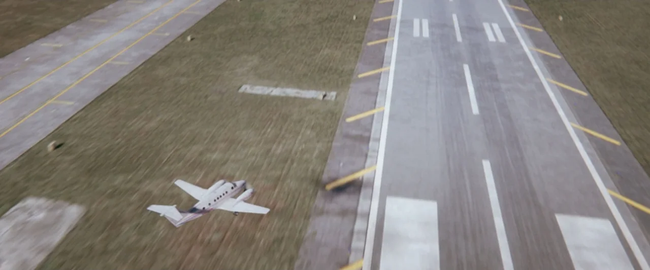 On a Wing and a Prayer (2023) movie still: establishing — An aerial view shows a small twin-engine aircraft flying over a concrete runway. Painted…
