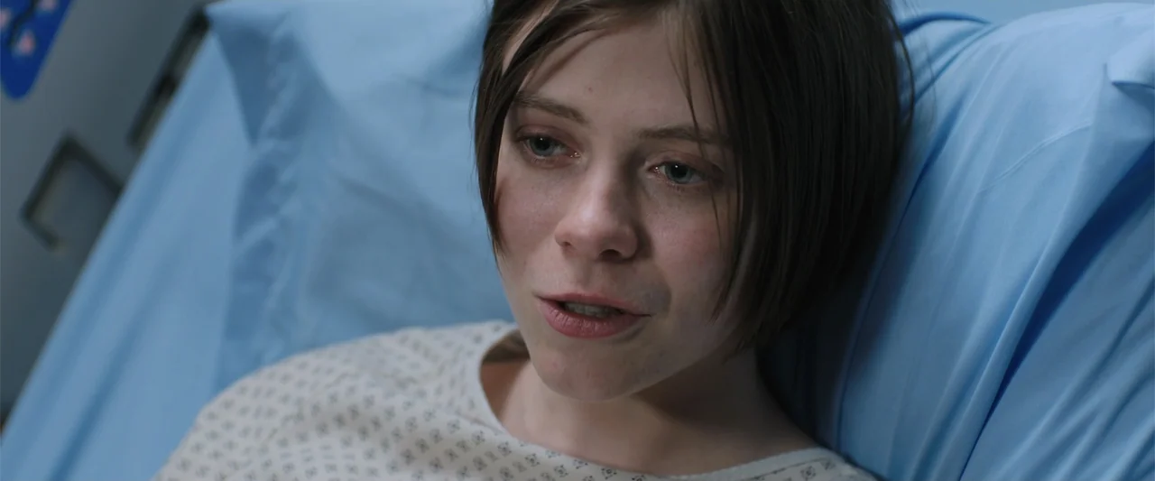 Trap House (2025) movie still: closeup — A sterile medical environment with light blue linens covering the bed. A young woman in a hospital gown…