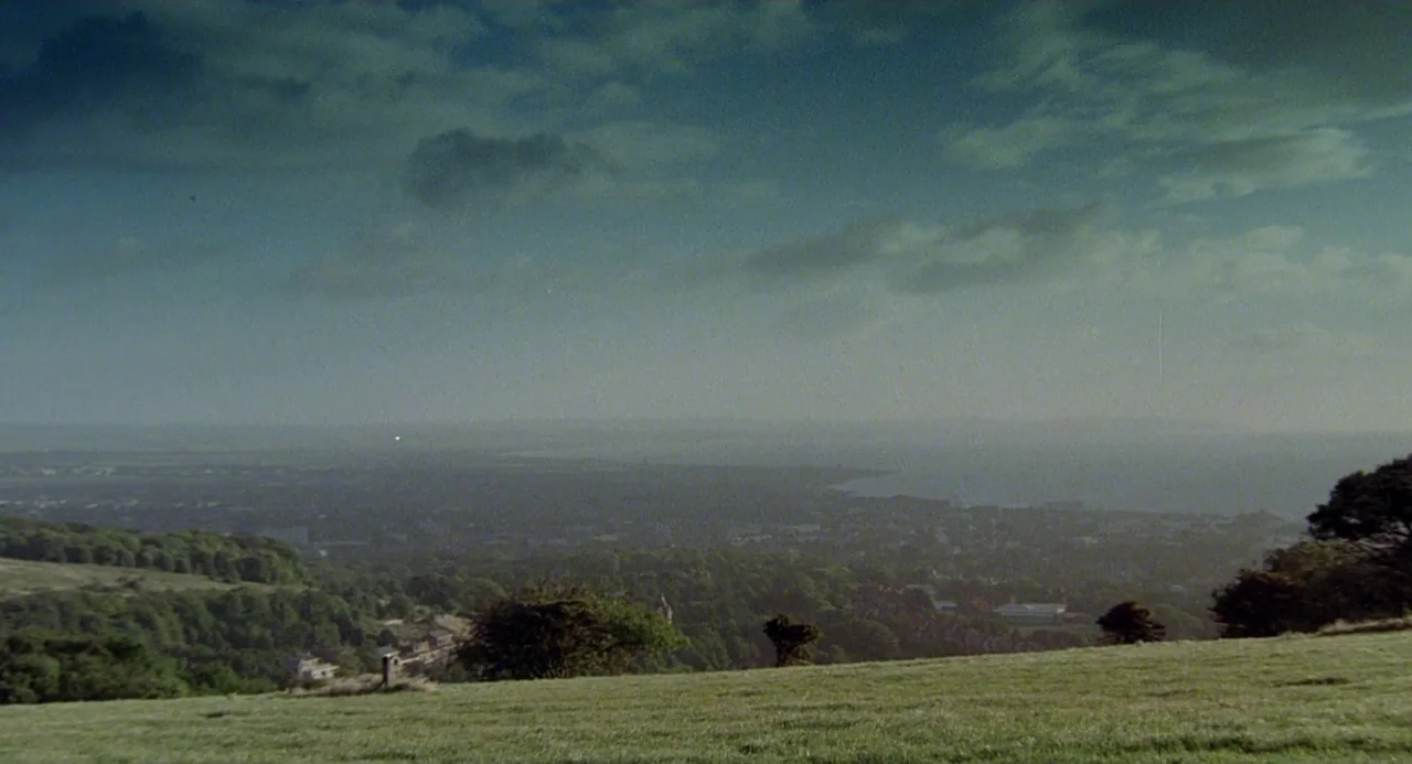 Quadrophenia (1979) movie still: establishing — A grassy hillside overlooks a vast valley area under a cloud-filled sky. Dense forest lines the…