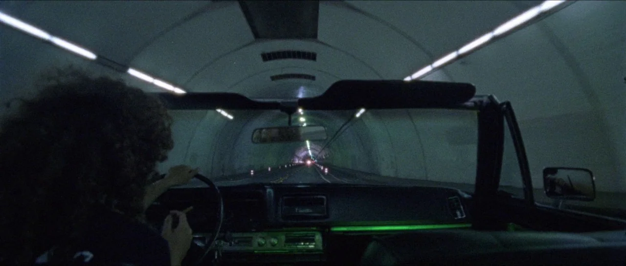 Bliss movie still: medium — A dark, curved tunnel is illuminated by rows of linear overhead lights. A person drives a convertible vehicle while…