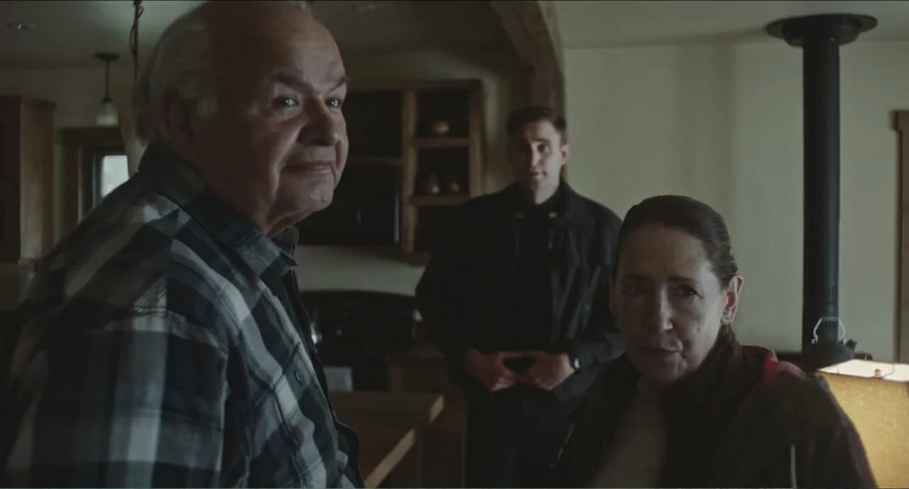 Cowboys (2014) movie still: medium — An interior living space with a dark wood structure and light-colored walls. Three individuals stand in a room,…
