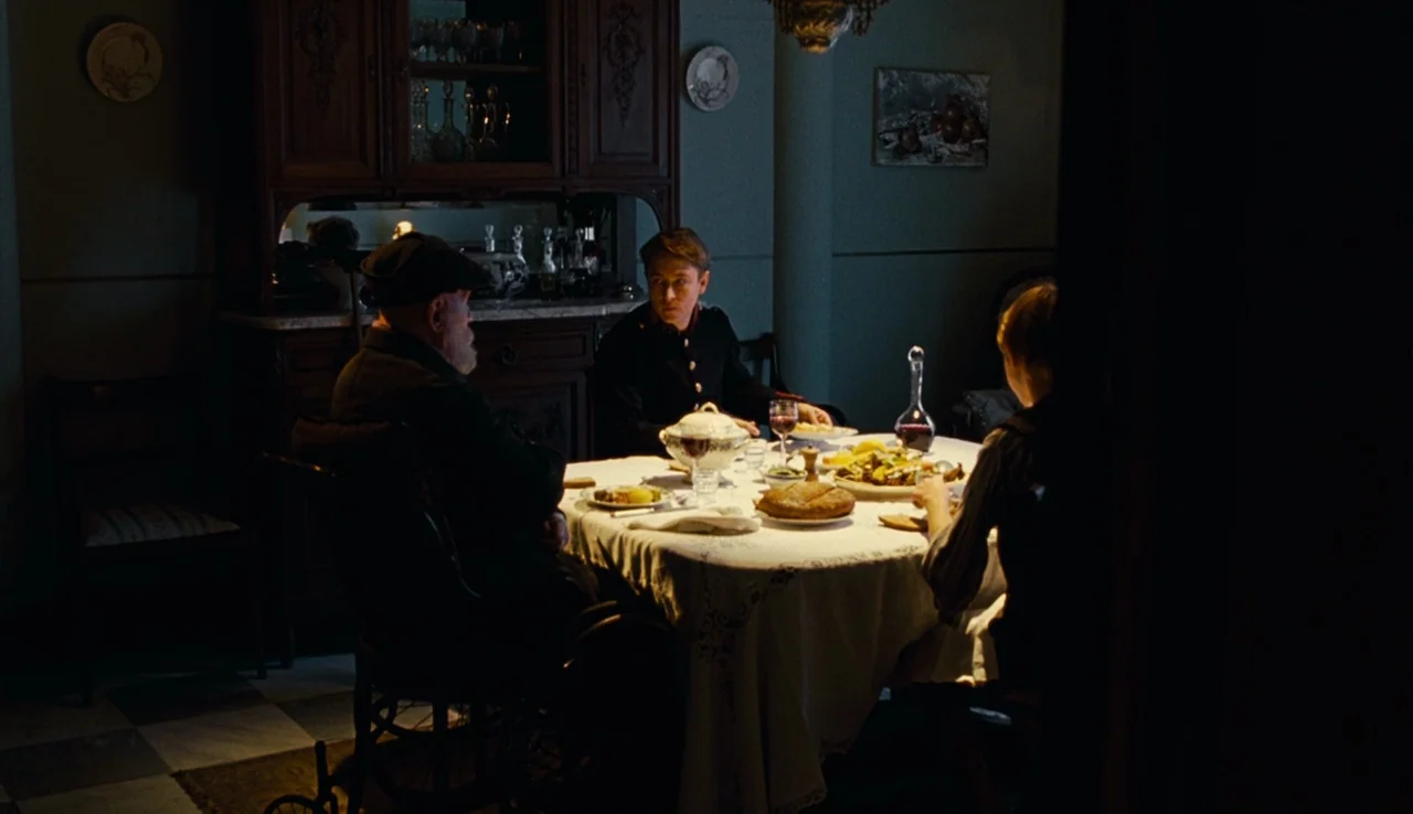 Renoir (2012) movie still: medium — A dimly lit room with a wooden hutch and a set dining table. Three people are seated around a tablecloth while…