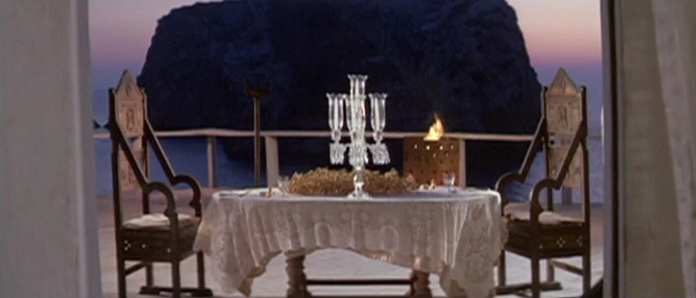 Boom! (1968) movie still: wide — An outdoor terrace overlooks the ocean with a large rock formation in the background. A table set for two with a…