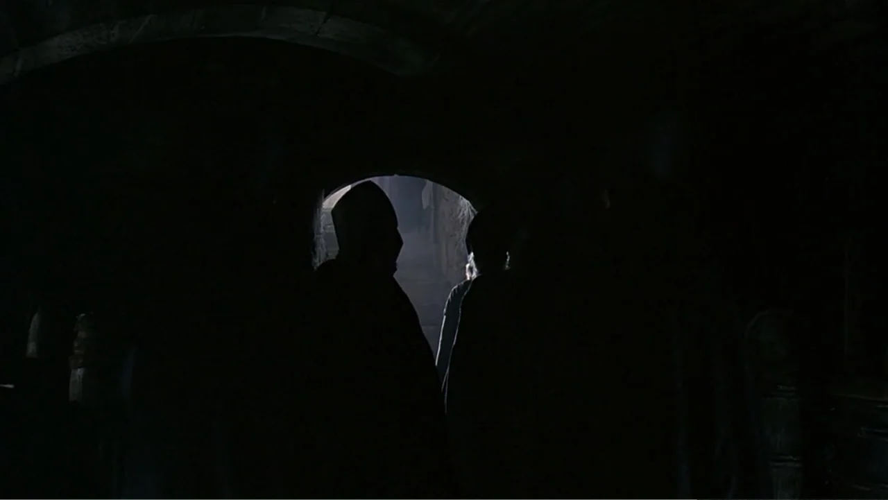 Elizabeth (1998) movie still: medium — Two silhouetted figures stand in a dark, arched stone passageway at night. Bright light illuminates a distant…