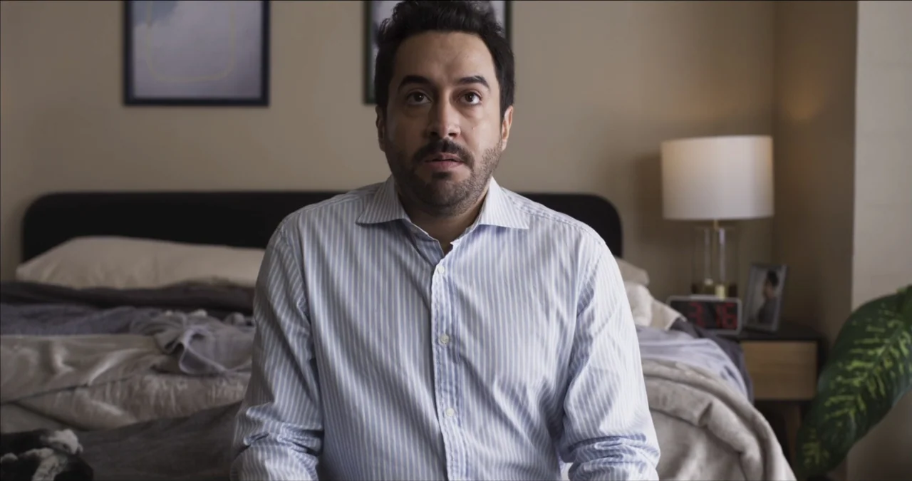 Bendita Suegra (2023) movie still: medium — A modern bedroom with a dark upholstered headboard and a wooden nightstand holding a lamp. A middle-aged…