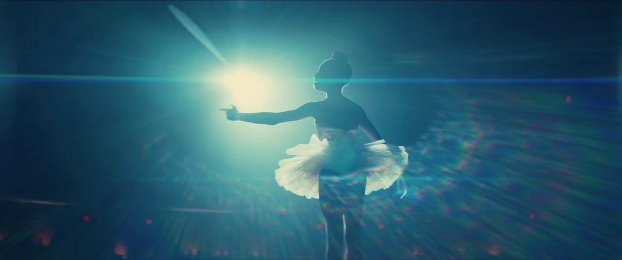 Us (2019) movie still: fullbody — A dark, expansive performance space with a single bright light focused behind a dancer. The ballerina stands…