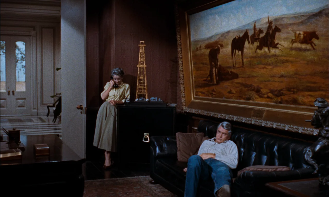 Giant (1956) movie still: wide — A dark-toned study features a large landscape painting on the wall and wooden furniture. A woman standing at a desk…