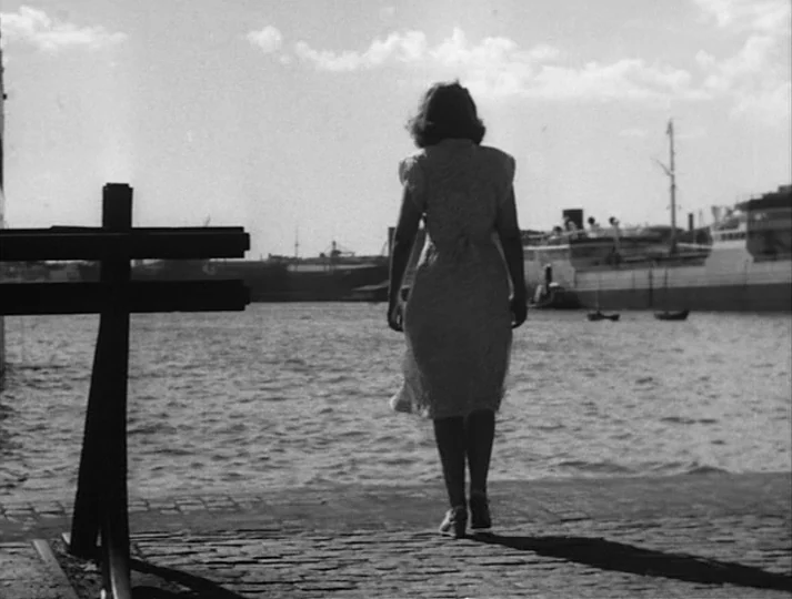 Port of Call (1948) movie still: fullbody — A stone-paved waterfront area overlooks a bay with large cargo ships in the distance. A woman walks…
