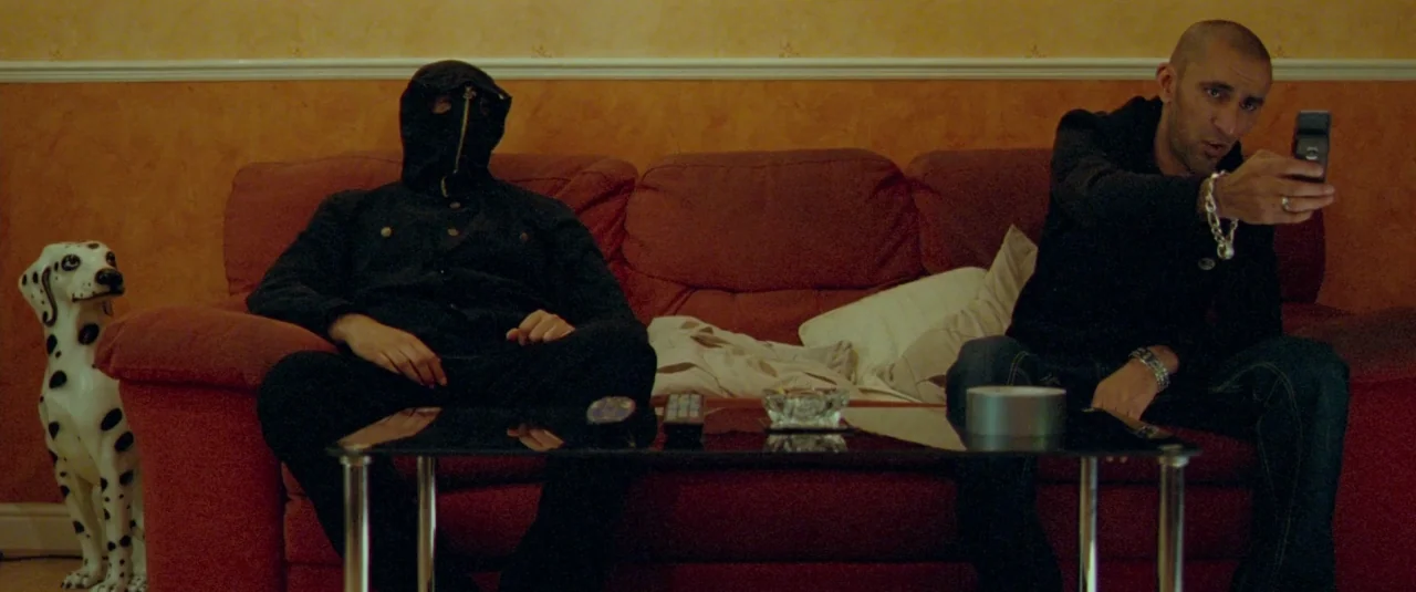 Catch Me Daddy (2014) movie still: medium — A domestic space features a red couch against warm orange walls and wallpaper trim. Two men sit on the…