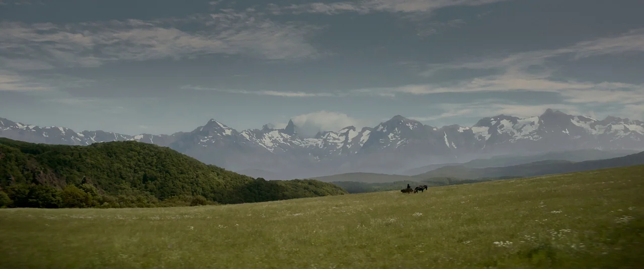 Ophelia (2018) movie still: wide — A vast, grassy mountain meadow stretches toward distant, snow-capped jagged peaks under a cloudy sky. A tiny…