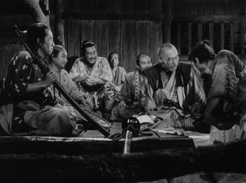 Seven Samurai (1954) movie still: medium — Seven men sit on wooden floorboards inside a dark timber structure with plank walls. The group discusses…