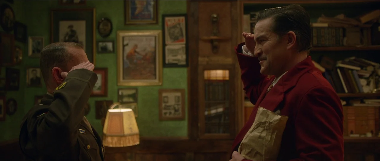 Brooklyn 45 (2023) movie still: medium — A study with green wallpaper covered in multiple small framed photographs and documents. A man in a red…