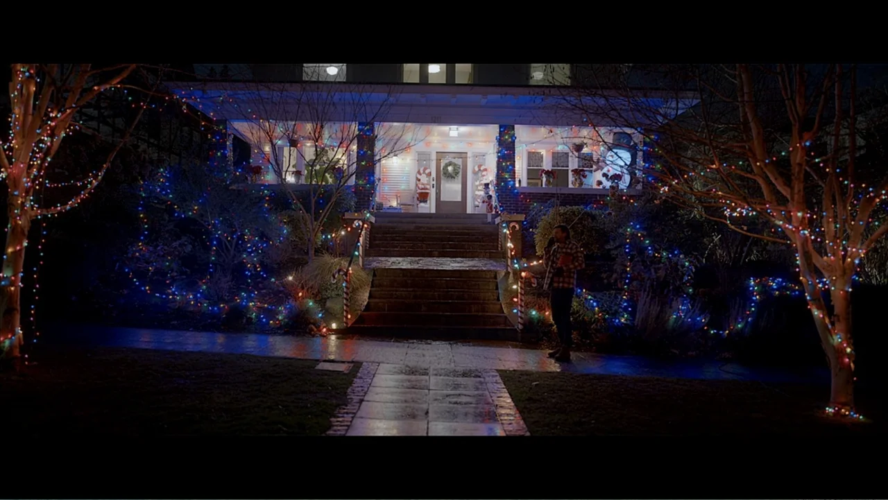 Jack in Time for Christmas (2024) movie still: wide — A dark residential house at night is covered in colorful strings of Christmas lights. A man…
