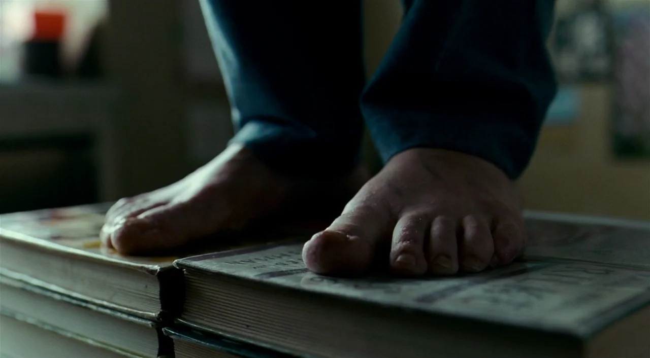 The Reader (2008) movie still: closeup — Two bare feet are placed firmly on the covers of two thick hardcover books. The person wearing dark…