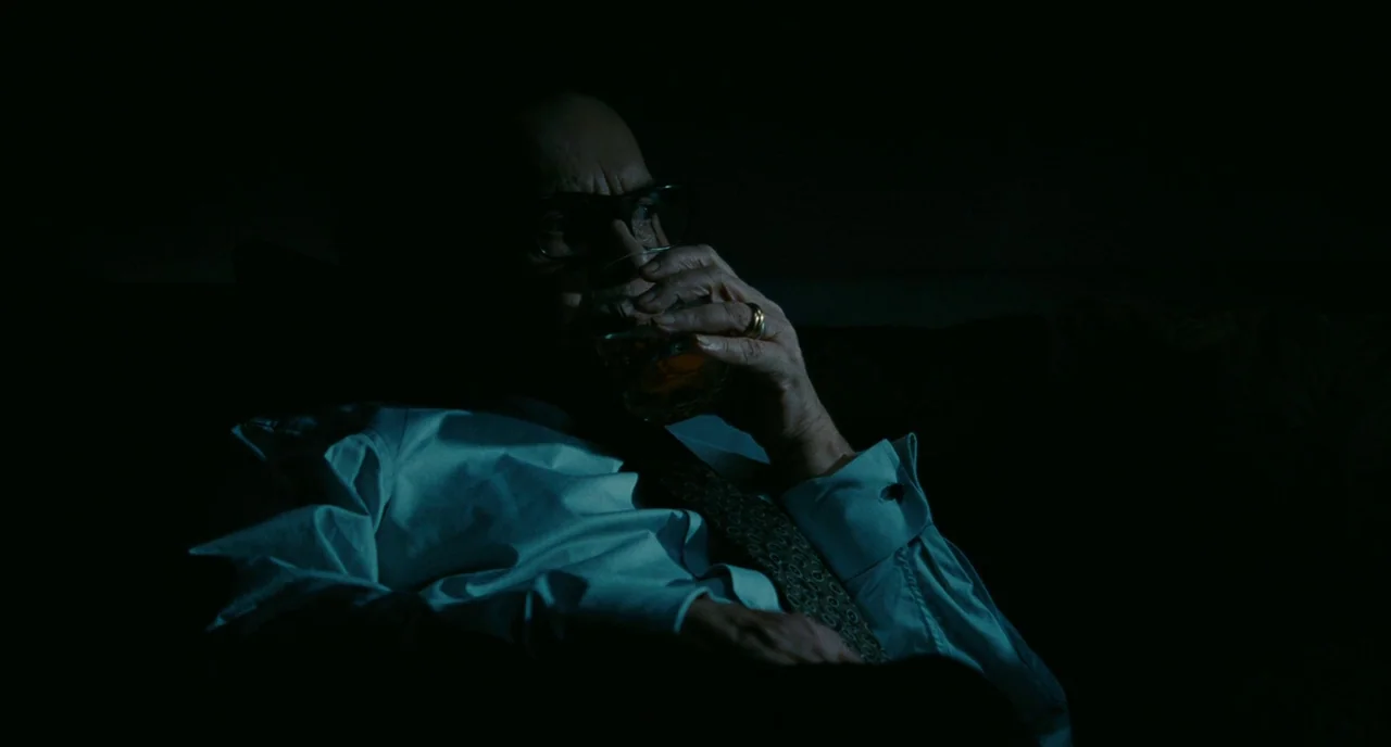 Saint Laurent (2014) movie still: medium — A man sits in darkness inside a room with minimal visible decor. He holds a glass of whiskey to his mouth.