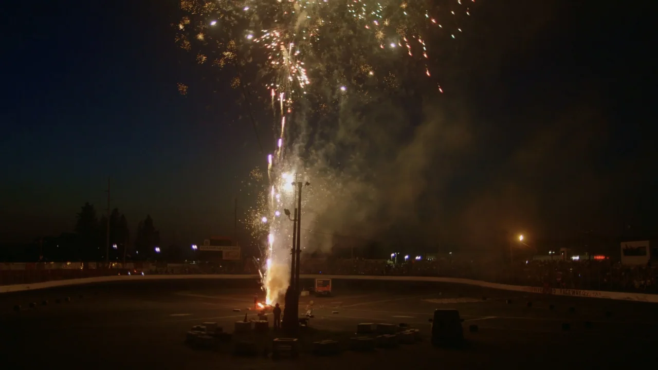 The Last Race (2022) movie still: wide — A wide view of an empty race track at night under a dark sky. Fireworks explode in the center of the track,…