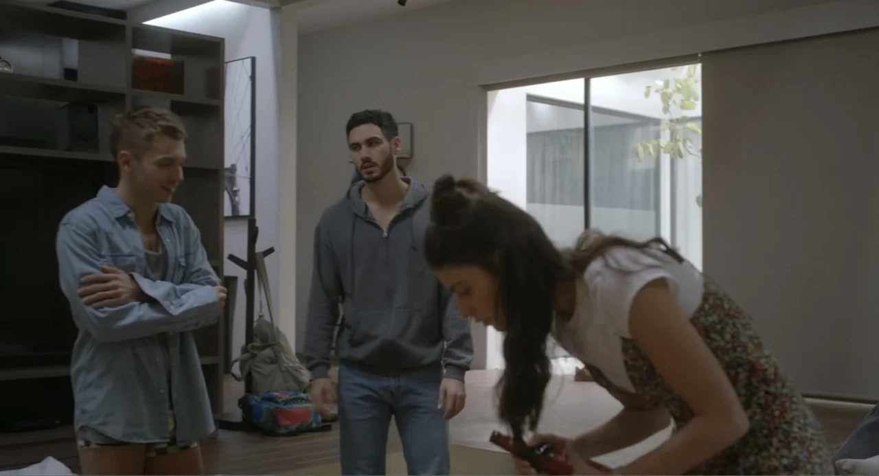 Straight (2024) movie still: medium — A modern apartment room with light walls and a large open window area. Three young adults interact in the…