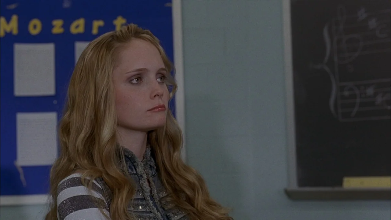 The Babysitters (2008) movie still: medium — A classroom interior with a blue display board and a chalkboard wall. A young woman with long blonde…