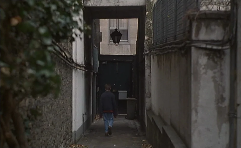 Kung Fu Master! (1988) movie still: wide — A narrow stone alleyway is bounded by high concrete walls and metal fencing. A young boy walks away from…