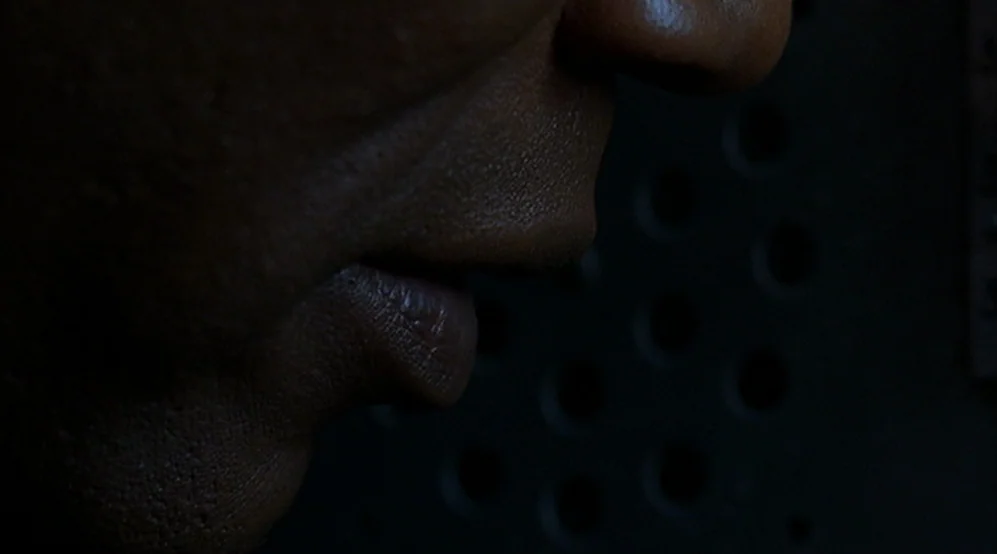 Jackie Brown movie still: closeup — A dark environment features a black metal panel with a grid of circular holes. A person's face appears in side…