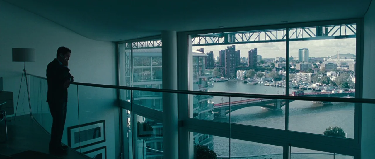 A Good Year (2006) movie still: wide — A modern apartment with a glass railing overlooks a city waterfront through floor-to-ceiling windows. A man…