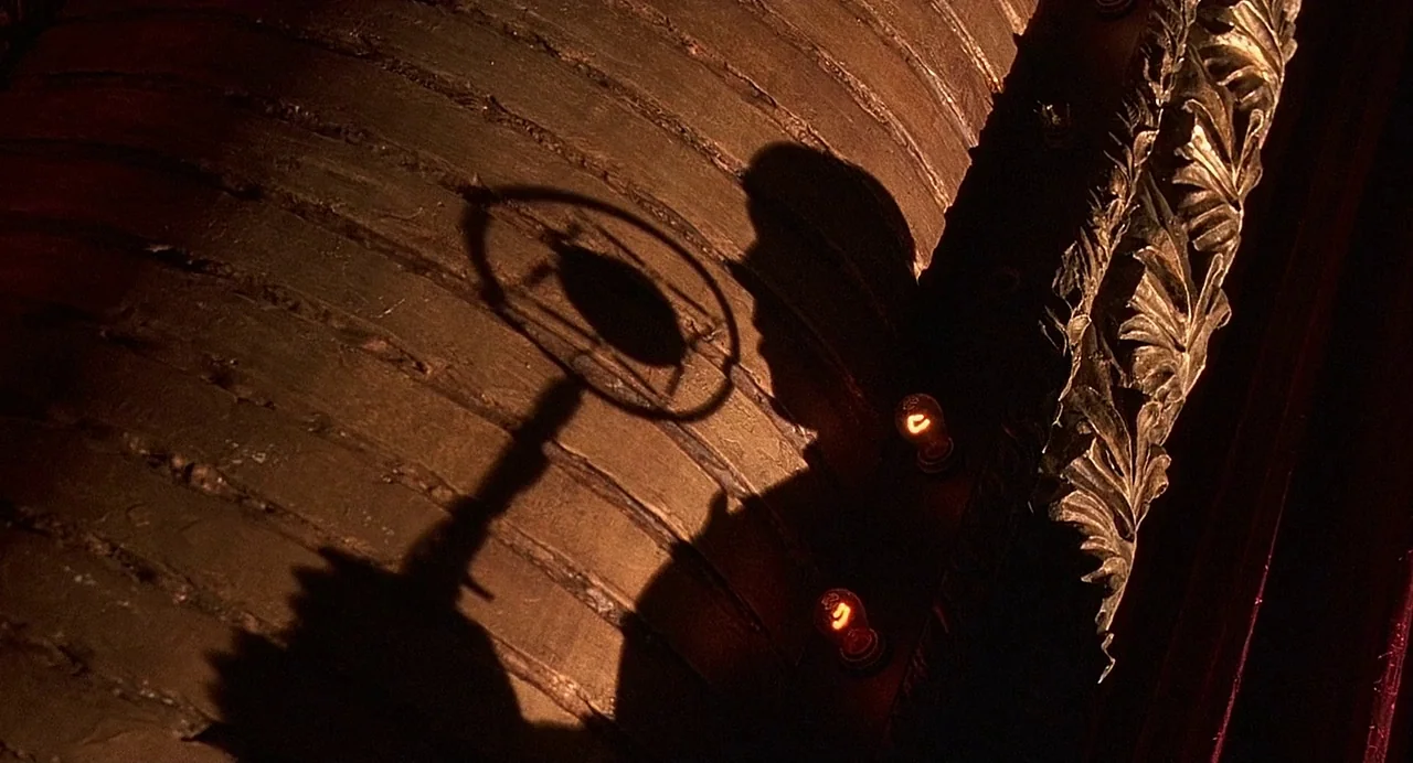 Chicago (2002) movie still: closeup — A dark shadow of a microphone and performer falls across a curved wooden panel. Ornate gold leaf molding and…