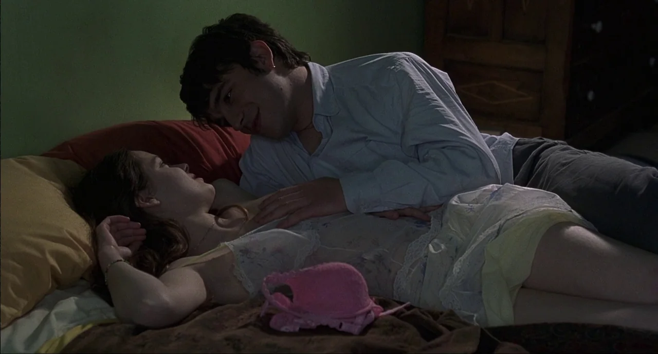 Fat Girl (2001) movie still: medium — A dimly lit room with green walls contains a couple lying on a bed. The man leans over the woman who lies on…