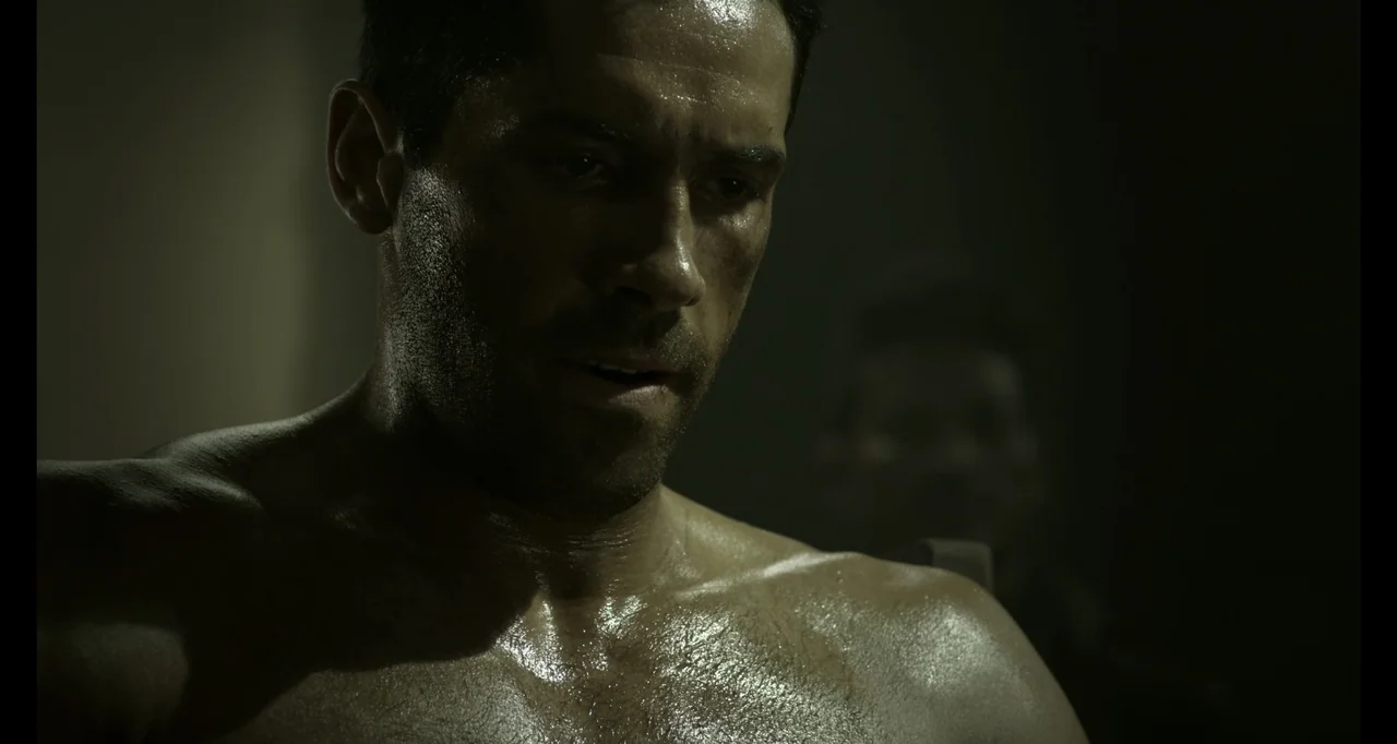 Ninja: Shadow of a Tear (2013) movie still: closeup — A dimly lit interior space with shadows obscuring the background. A muscular man with a sweaty…